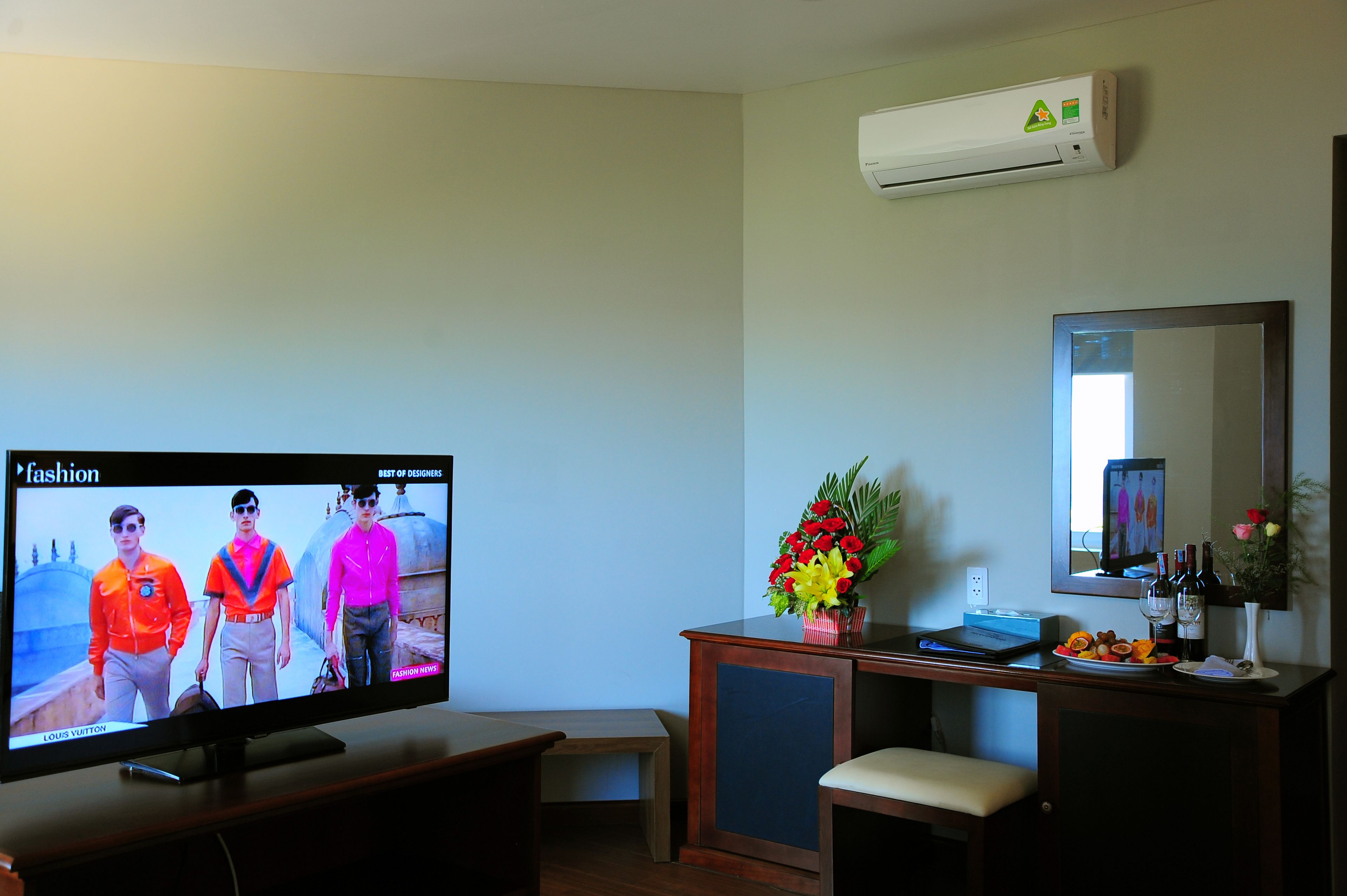 Una sala de estar con un televisor de pantalla plana mostrando un desfile de moda, un escritorio de madera con un jarrón de flores y un espejo en la pared.