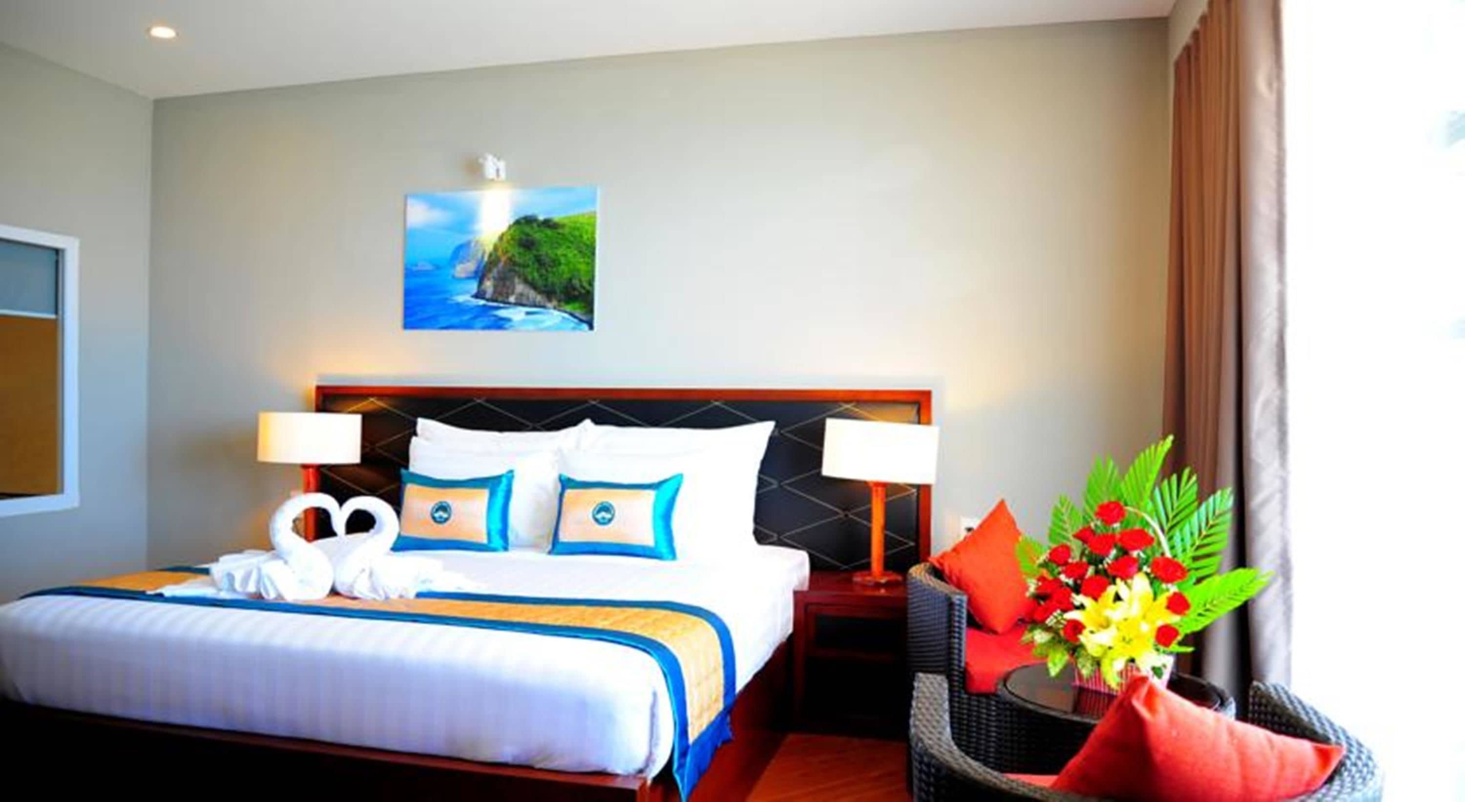 Una habitación de hotel con cama, dos lámparas de noche, un cuadro en la pared, una ventana con cortinas y una pequeña mesa con un arreglo floral.