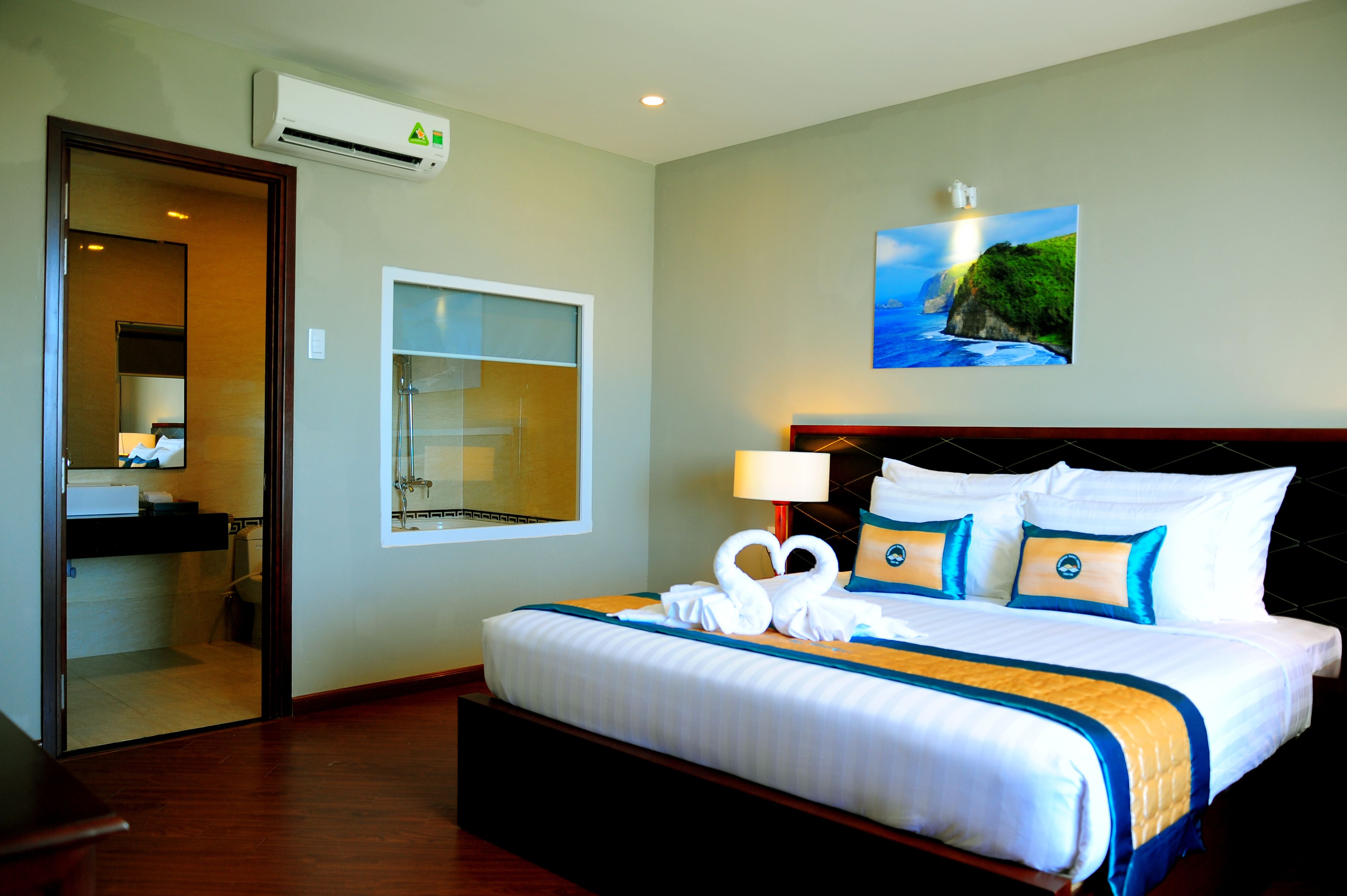 A hotel room with a large bed, a bathroom visible through an open door, a wall-mounted air conditioner, and a painting of a coastal scene.