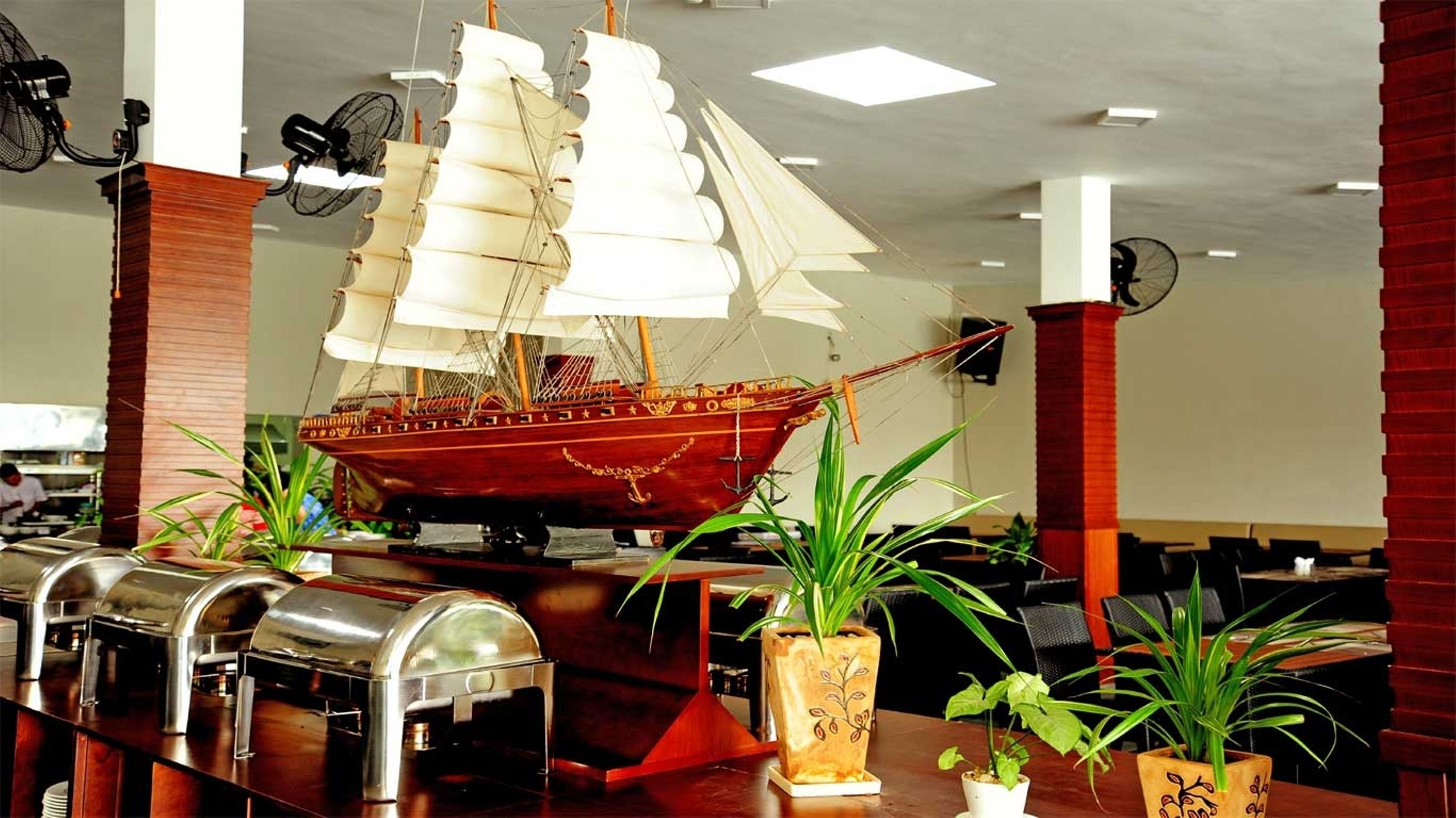 A ship model displayed in a lobby area with buffet counters and potted plants.