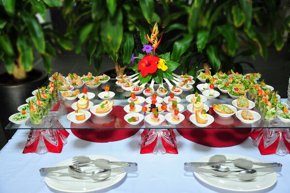 Una mesa de buffet con diversos alimentos dispuestos en platos y copas blancas, sobre un fondo de plantas verdes.