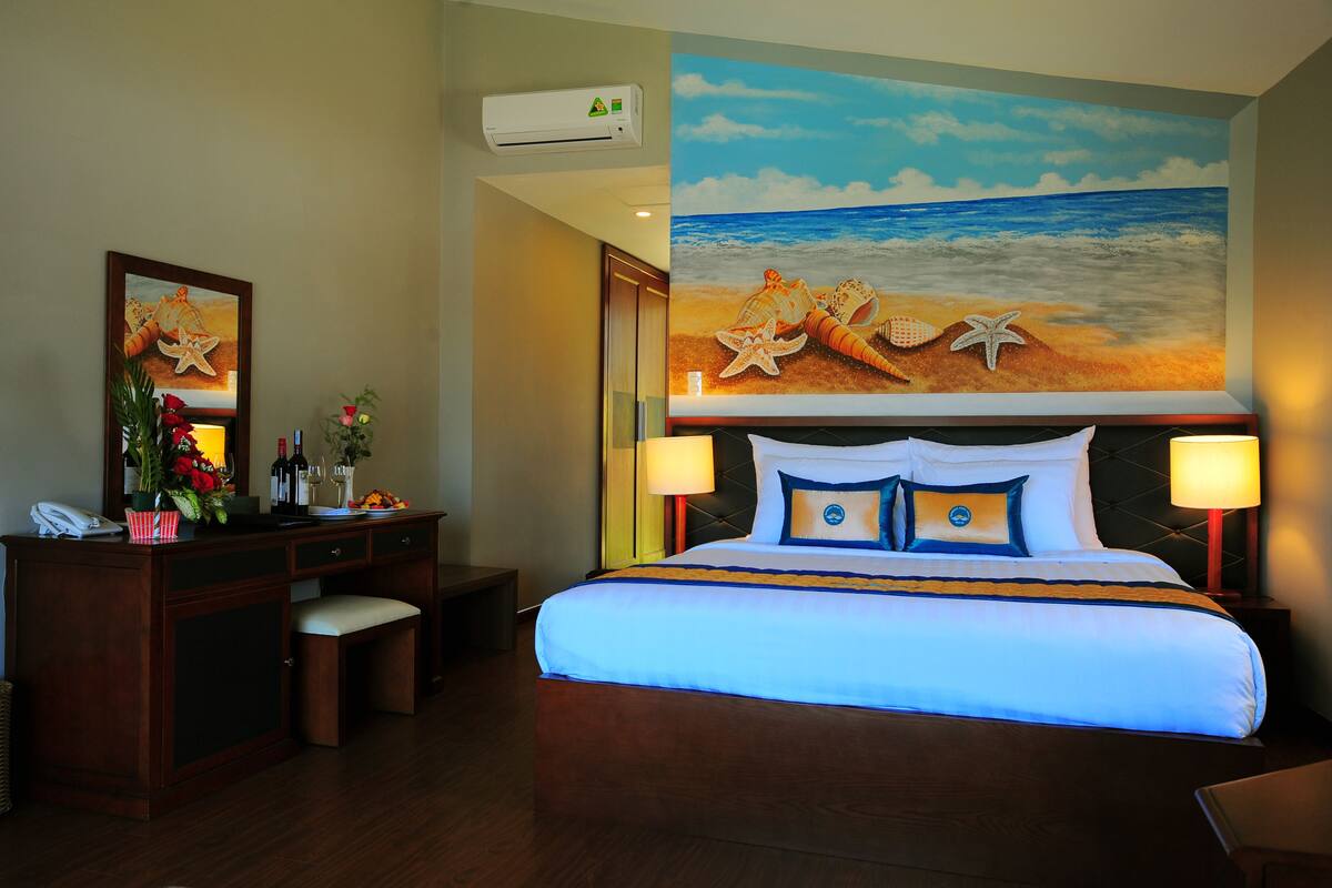 A hotel room with a bed, a desk, a chair, a dresser, and a large beach-themed mural on the wall.