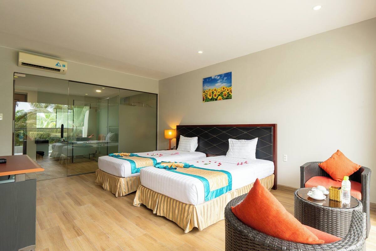 A hotel room with two beds, a sitting area with a wicker chair, a glass-enclosed shower area, and a painting on the wall.