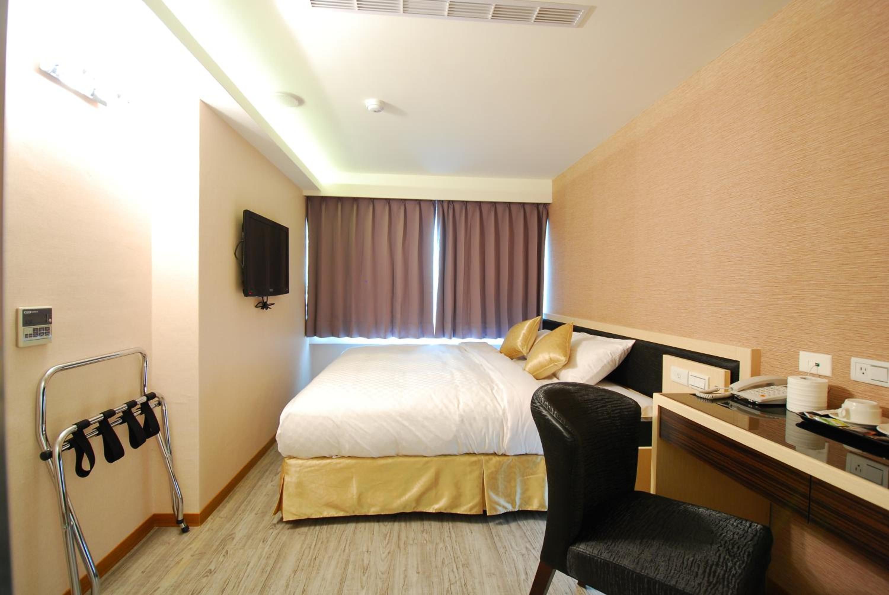 A hotel room with a bed, a desk with a chair, a television, a bedside table with a phone and a lamp, and a handrail.