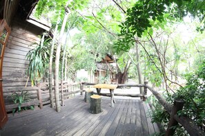 A wooden deck with a table and stools, surrounded by trees and a rustic building. - Natural Bungalows Restaurant and Bar (Kampot)