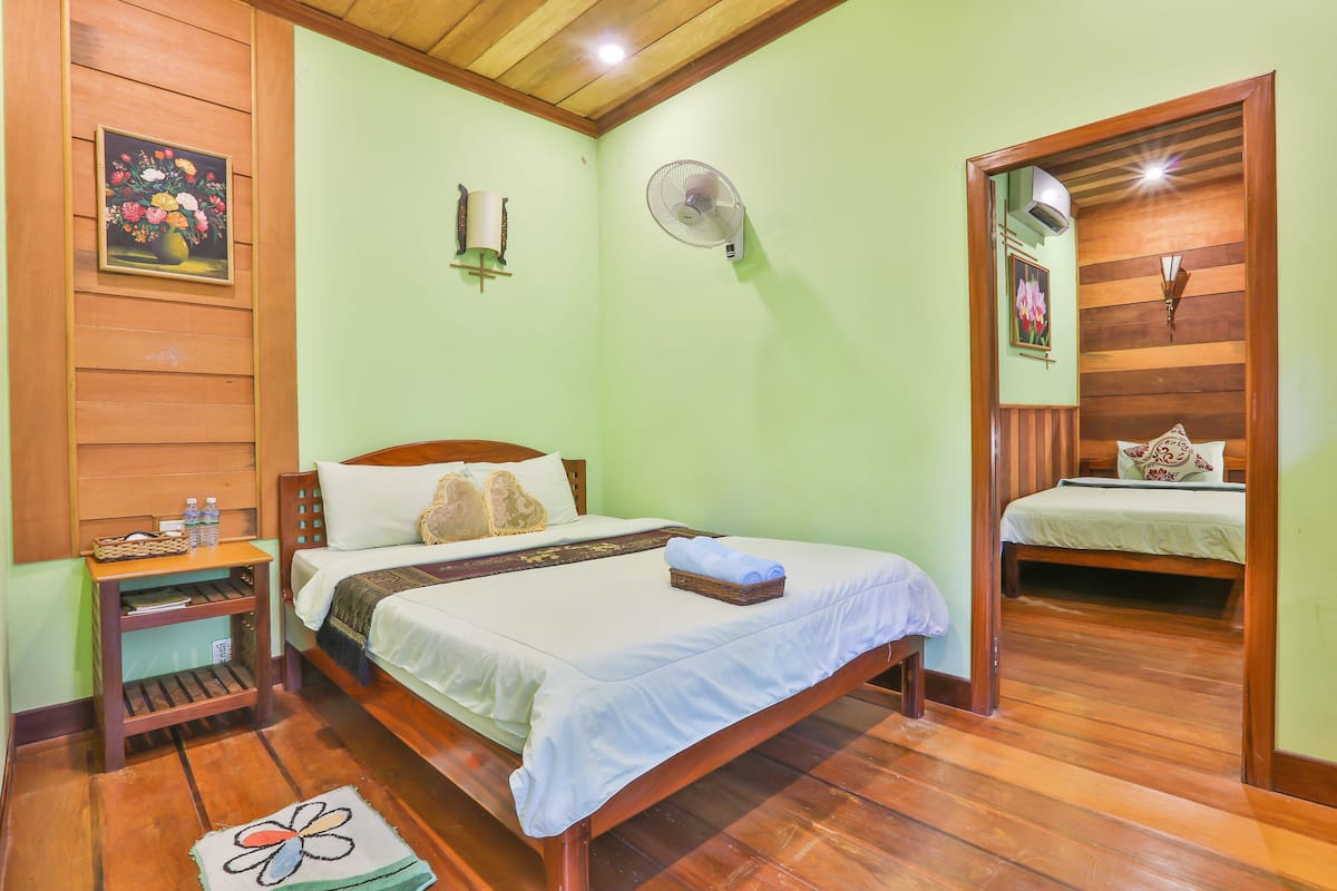 A room with a wooden bed, a nightstand, a fan, and a painting on the wall.