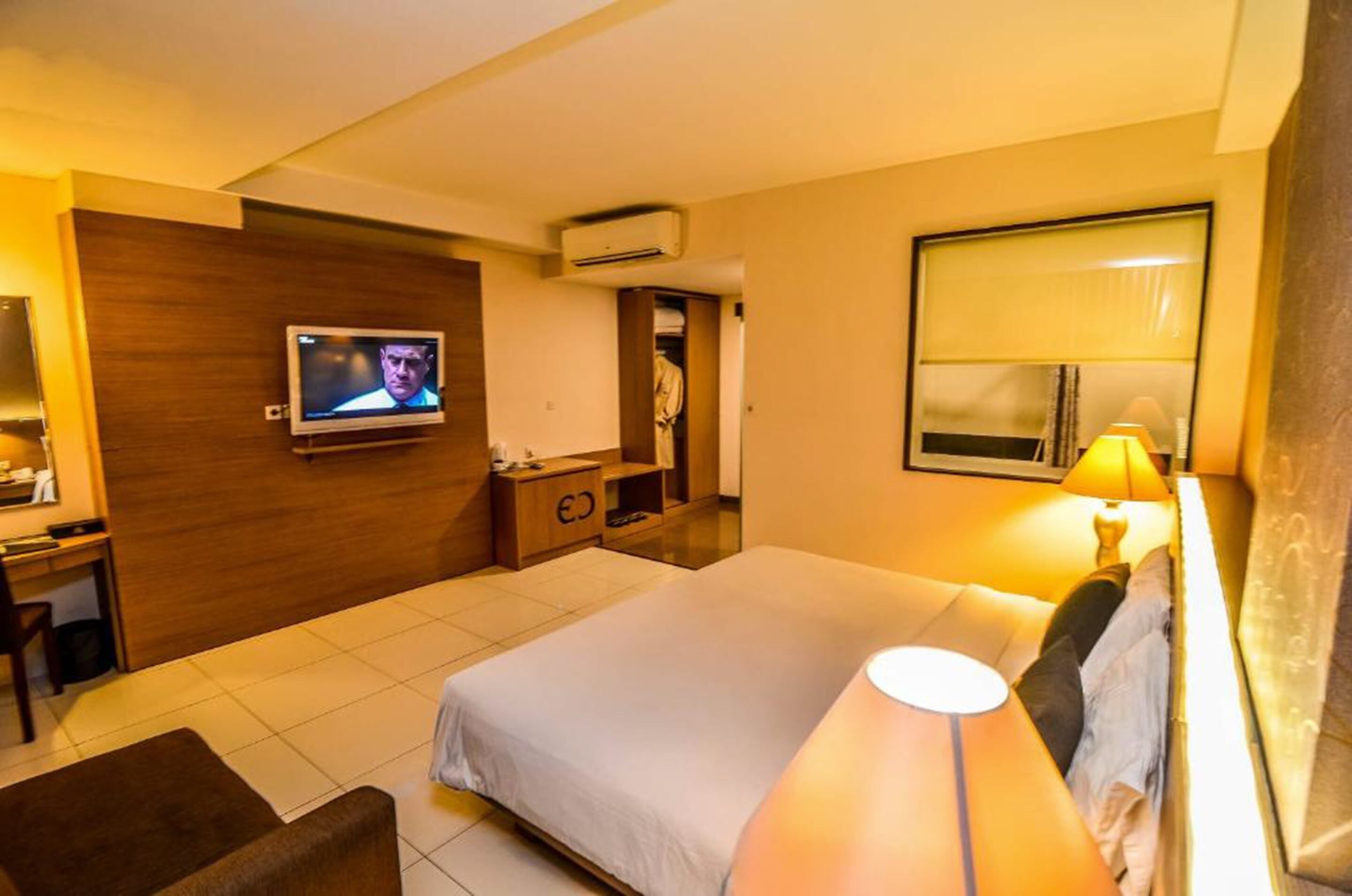 A hotel room with a bed, a TV mounted on the wall, a desk, a chair, and a bedside table.