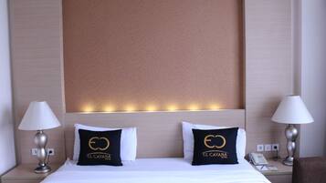 Quarto de hotel com uma cama grande, duas almofadas com o logotipo do hotel, luminárias de cabeceira e mesa de cabeceira com um telefone.