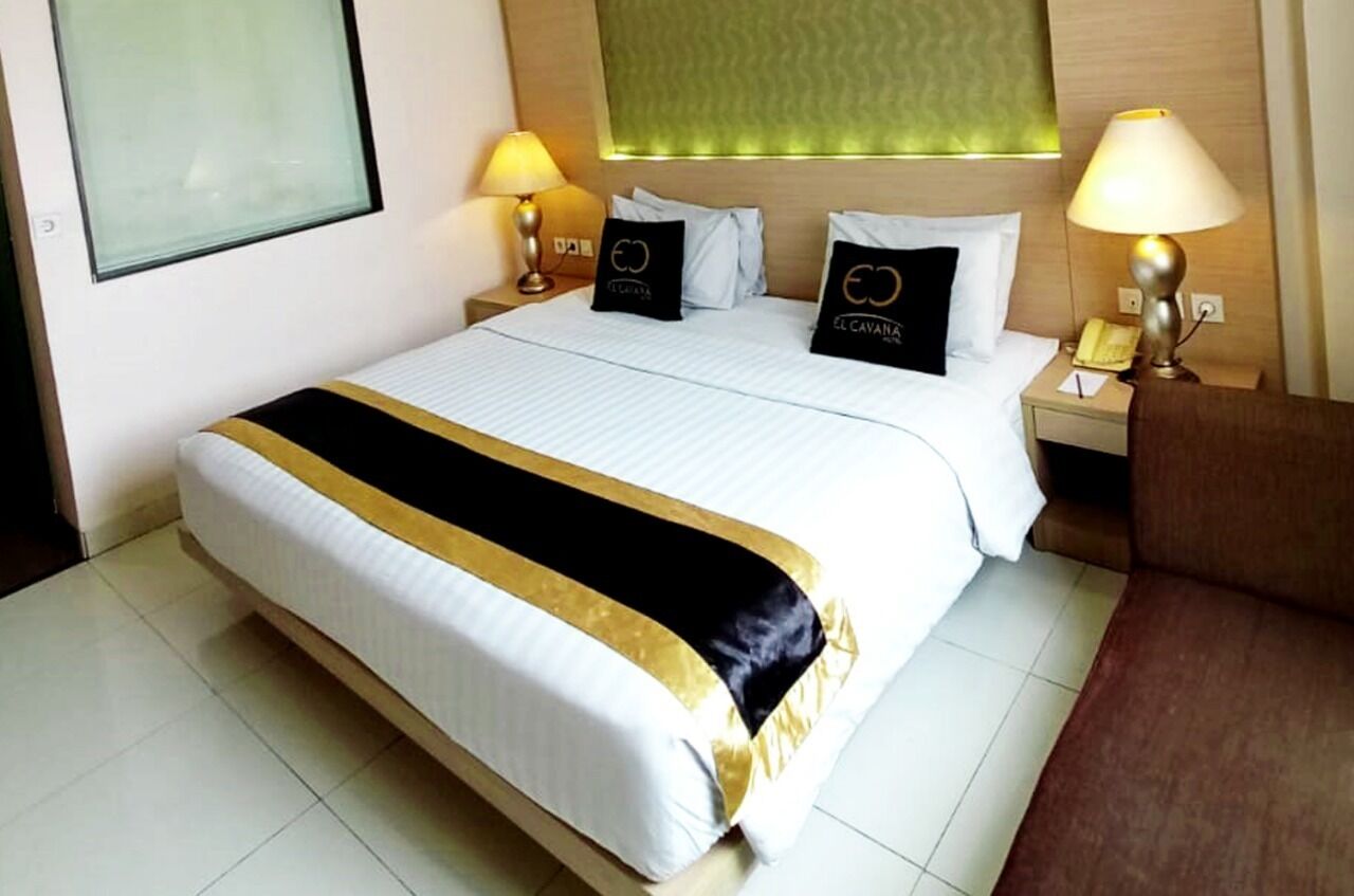 A hotel room with a large bed, two bedside tables with lamps, a headboard with a green panel, and a brown armchair.