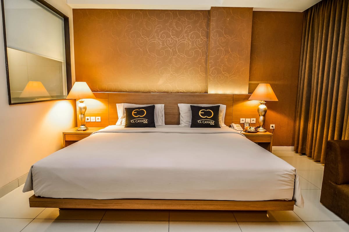 A hotel room with a large bed, two bedside lamps, a nightstand with a phone, and a headboard with a patterned design.