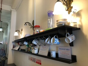 A breakfast area with a shelf holding mugs, a water bottle, and a sign indicating breakfast options. - Pillow Talk Hostel (Singapore)