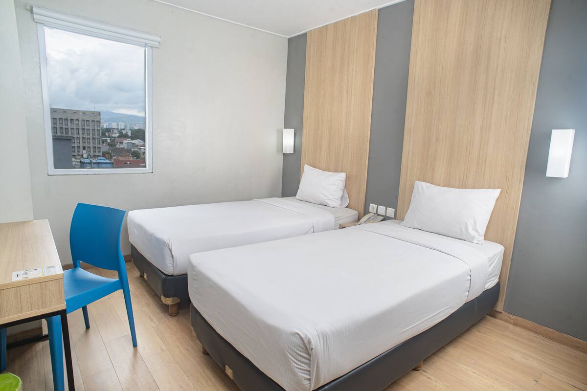 A hotel room with two single beds, a desk, a chair, and a window with a city view.