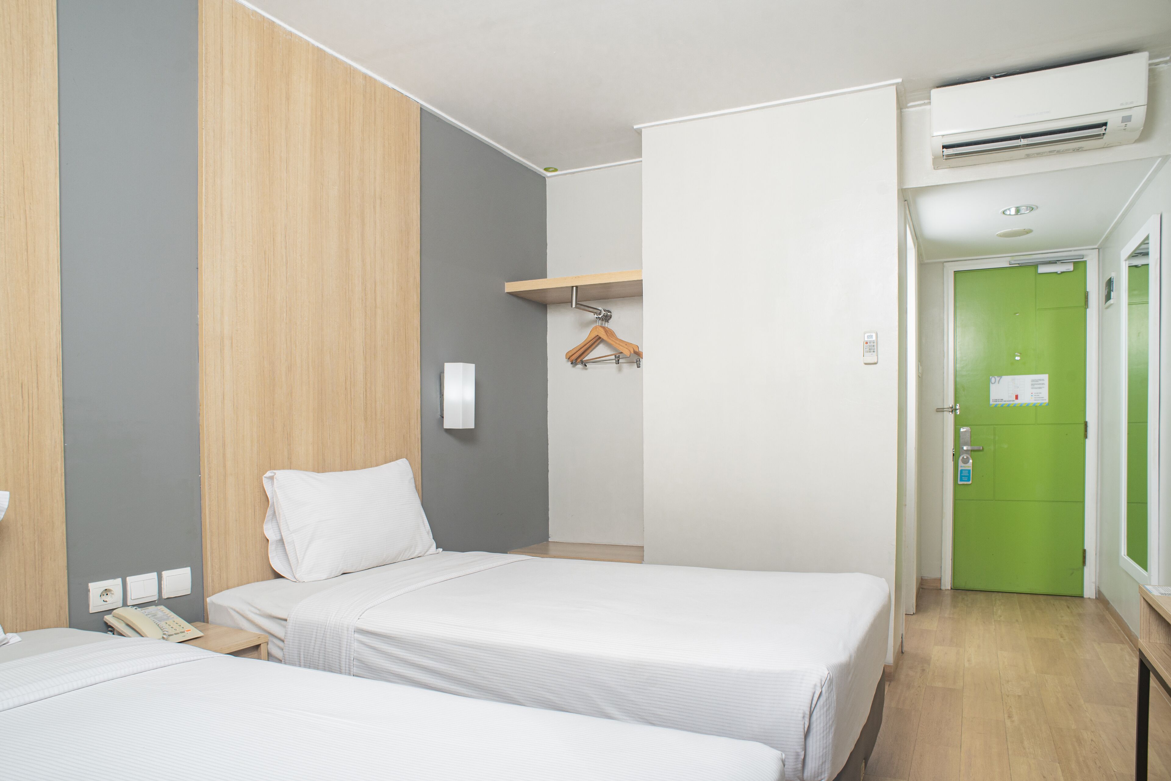 A hotel room with two beds, a wooden panel wall, a green door, and a small table with a telephone.