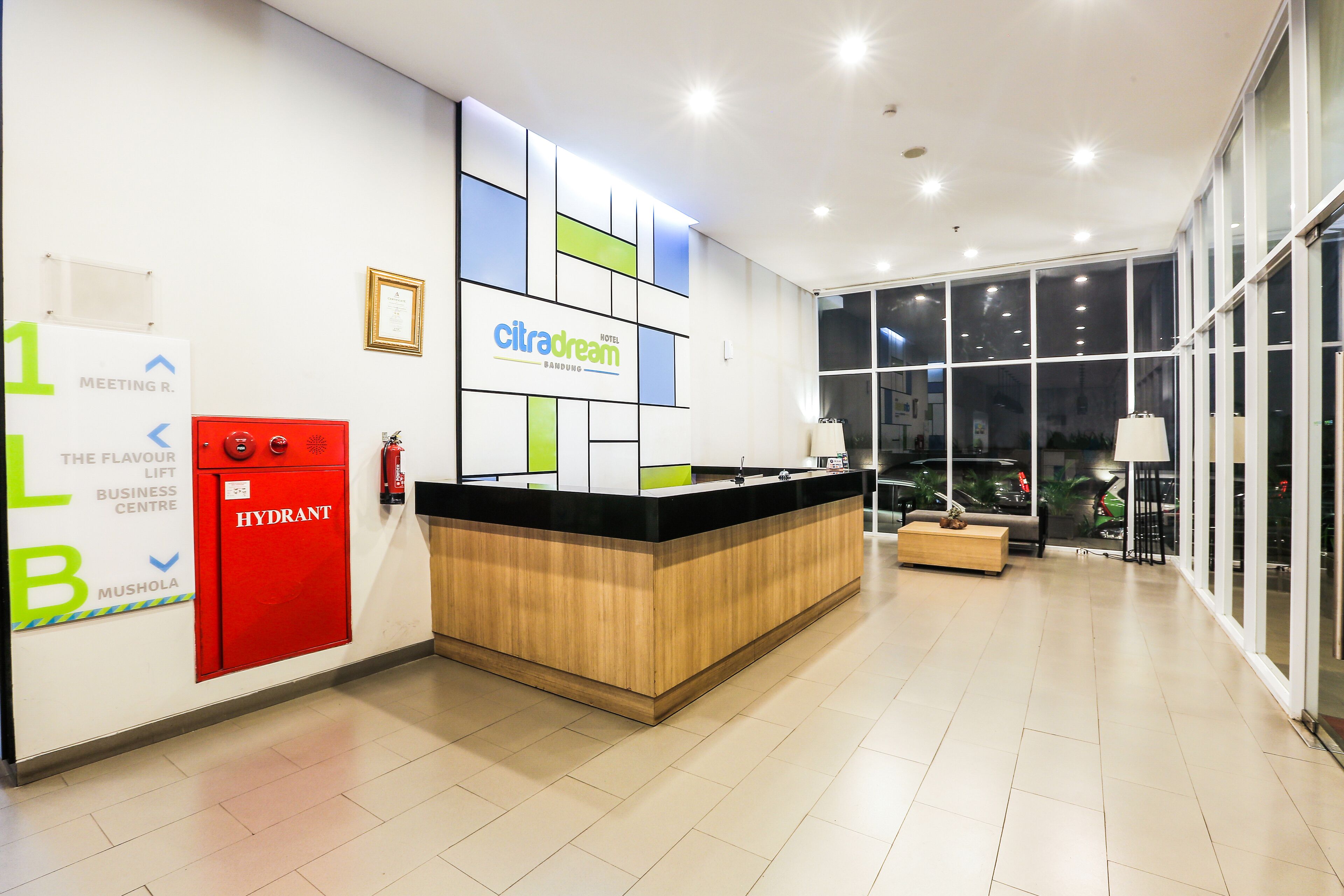A hotel reception area with a modern design, including a reception desk, a fire extinguisher, and a large window offering a view of the outside.