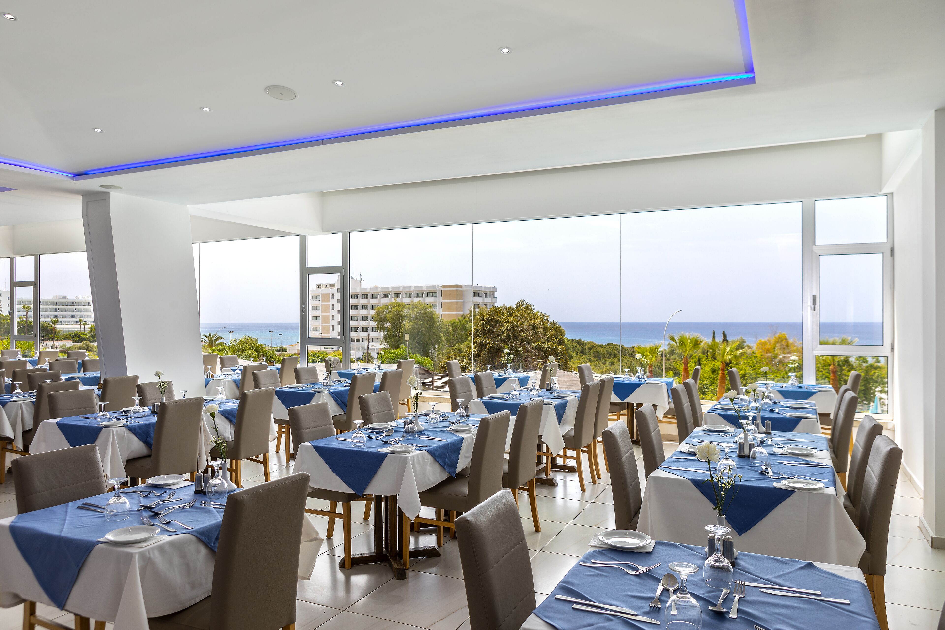 Un restaurant où les tables sont dressées pour le service, de grandes fenêtres offrant une vue sur la mer et un décor intérieur moderne.