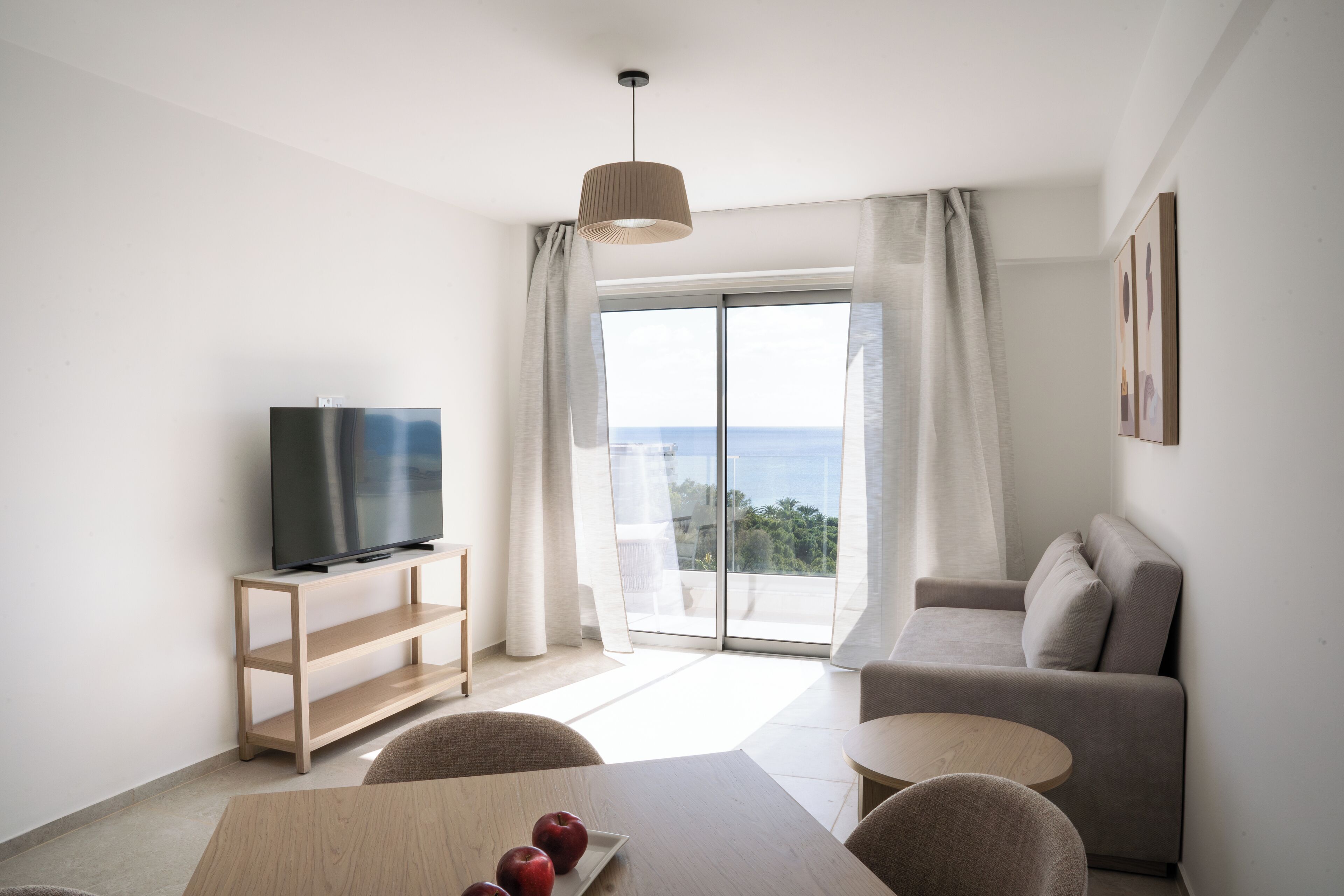 Una sala de estar moderna con un televisor de pantalla plana sobre un mueble de madera, un sofá, una mesa de comedor con manzanas y un ventanal con vista al mar.