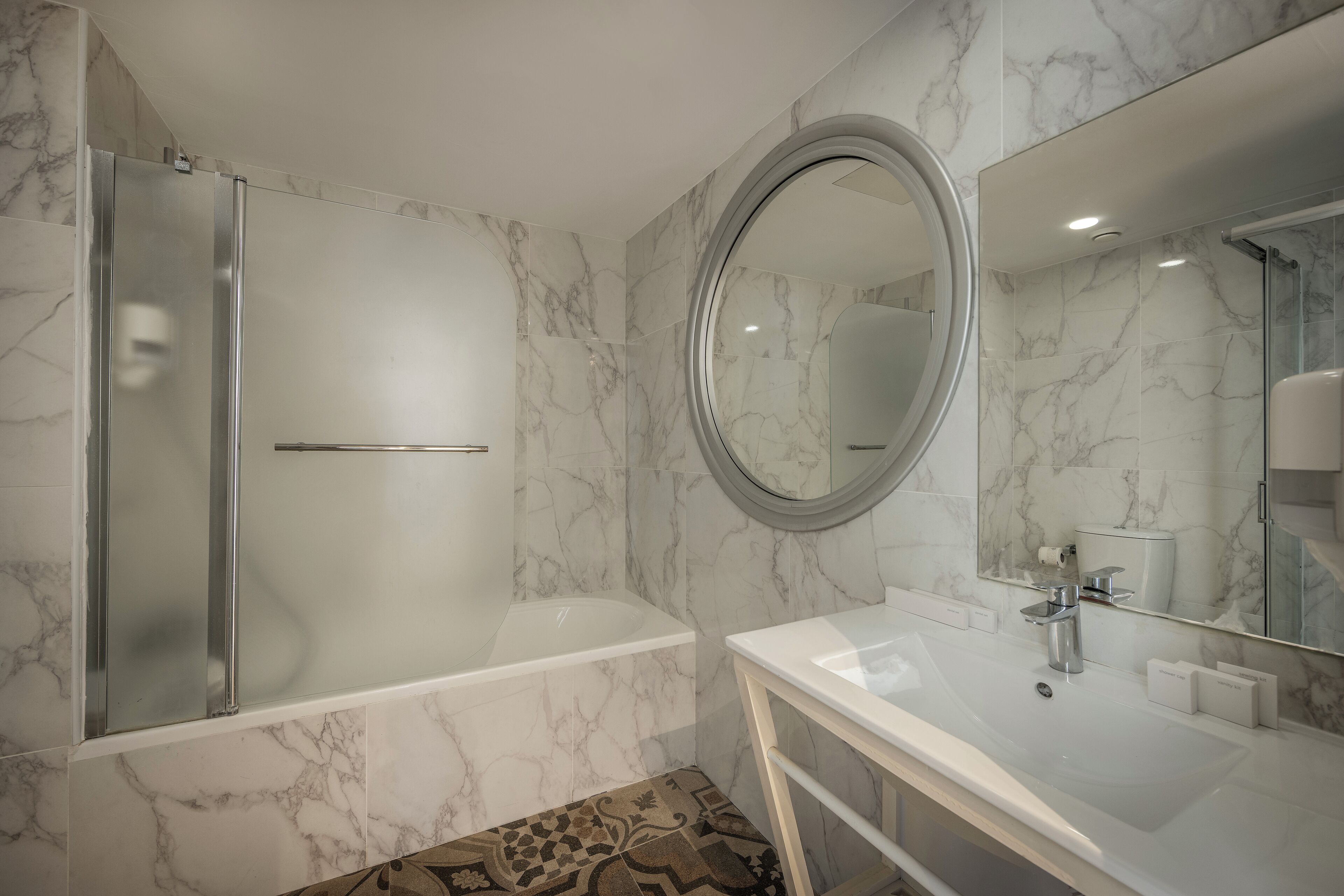 A modern bathroom with a marble-tiled wall, a large round mirror, a bathtub, and a white sink.