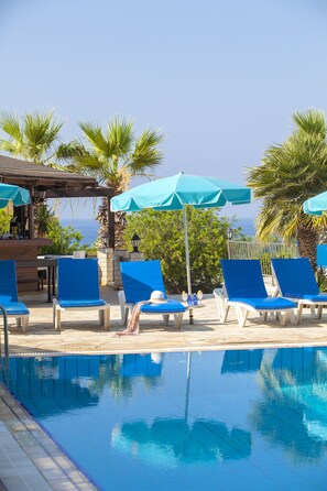 Ein Poolbereich mit blauen Liegestühlen und Sonnenschirmen, eine Bar sowie der Blick auf den Ozean.