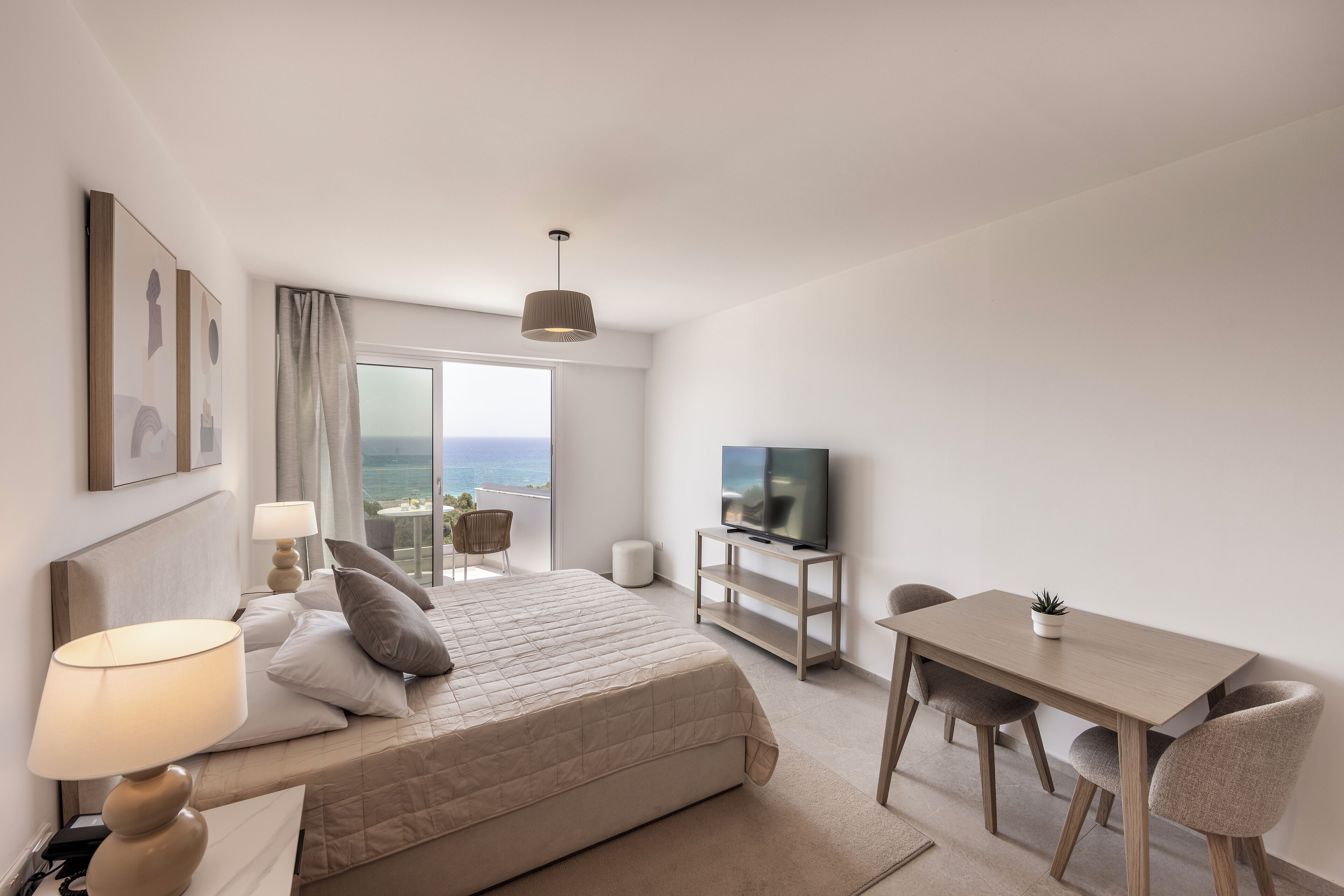 Een moderne hotelkamer met een bed, nachtlampjes, een televisie, een eettafel en uitzicht op zee.