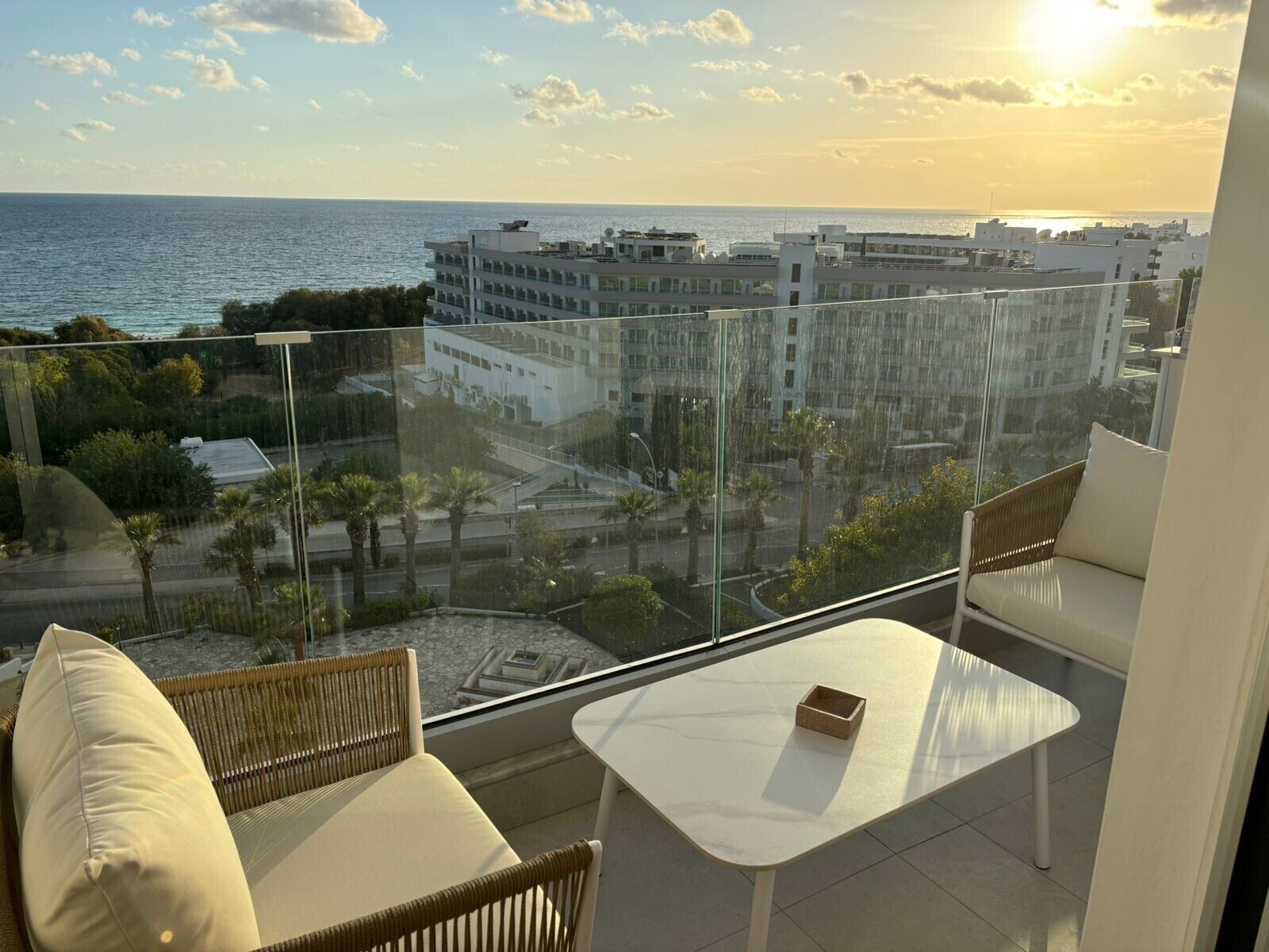 A balcony with a view of the sea and buildings. There is a white table and two wicker chairs.