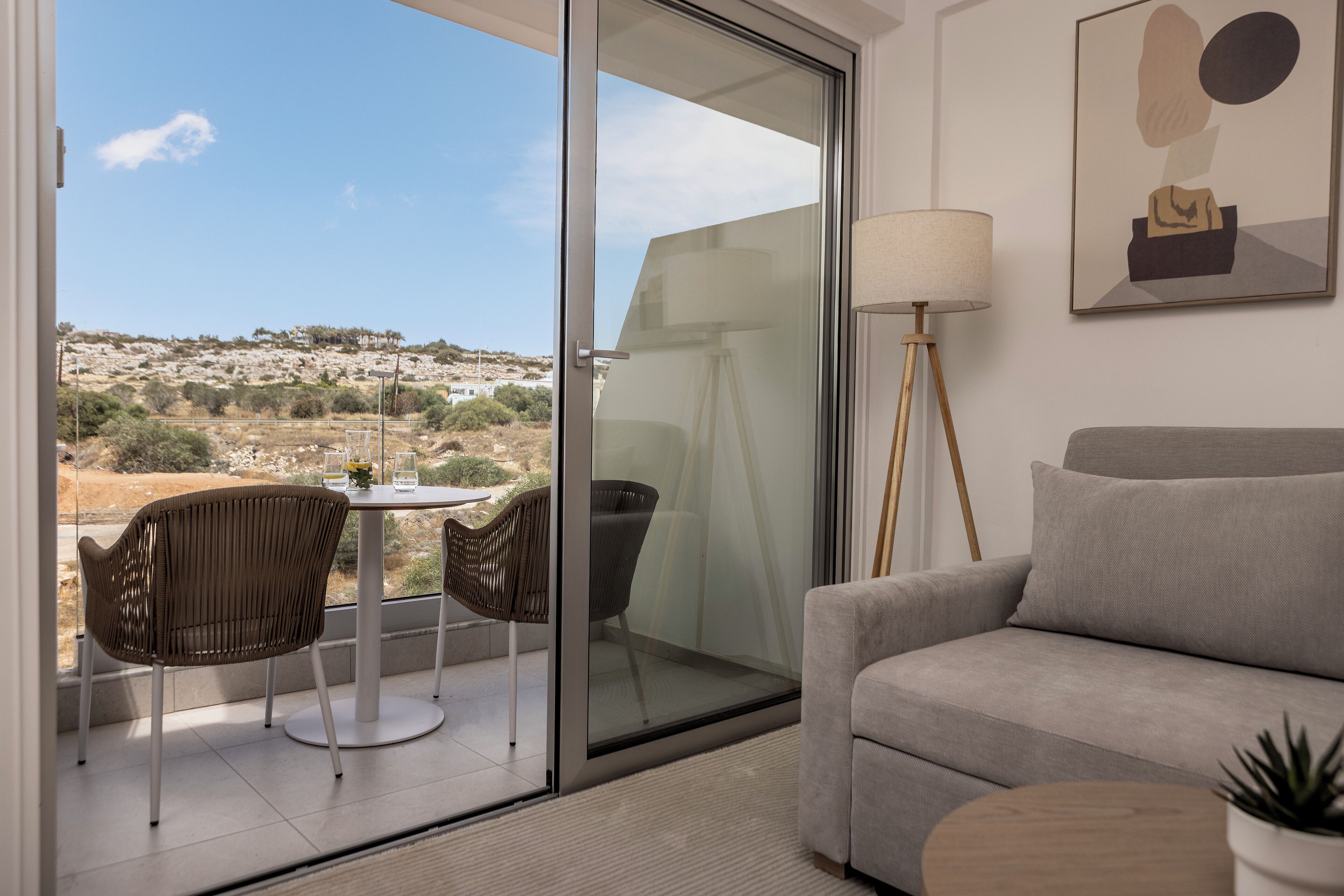 A modern living room with a sofa, a dining table with chairs, a floor lamp, and a view of the countryside through a sliding glass door.
