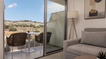 A modern living room with a sofa, a dining table with chairs, a floor lamp, and a view of the countryside through a sliding glass door.