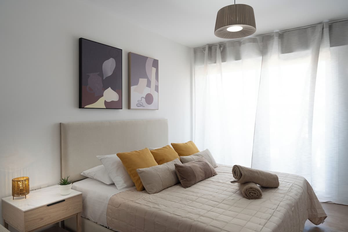 A neatly made bed with a beige quilt, a bedside lamp, and a rolled towel on the bed.