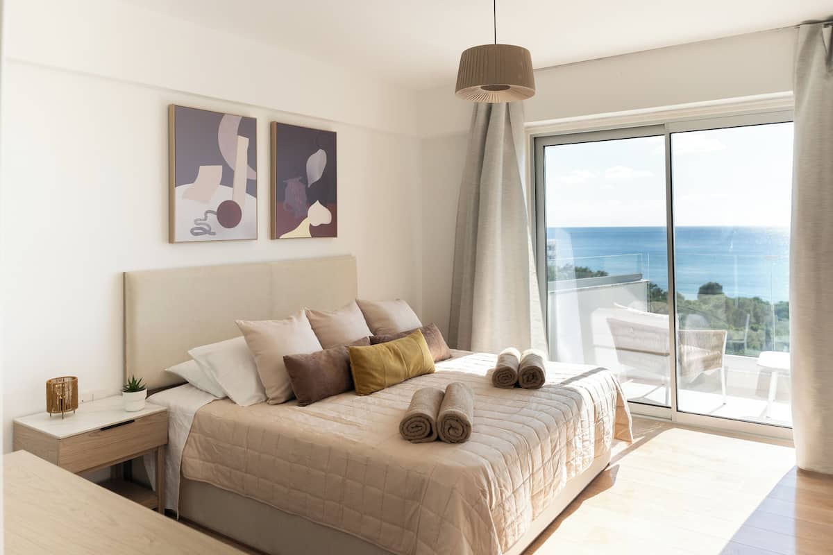 A modern bedroom with a large bed, a bedside table, a hanging light, and a view of the sea.