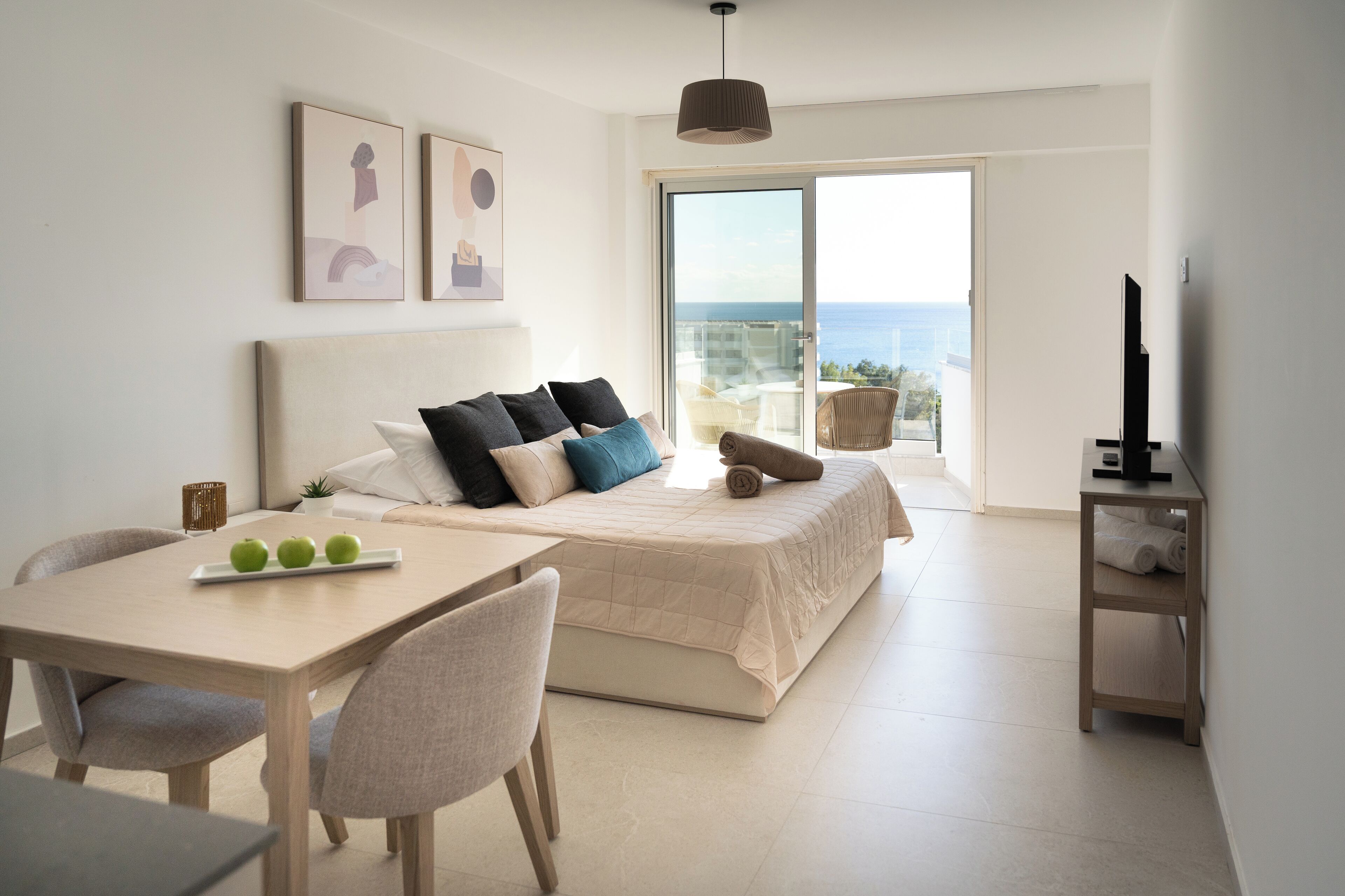 A modern bedroom with a bed, a dining table, and chairs. There is a balcony with a view of the sea.