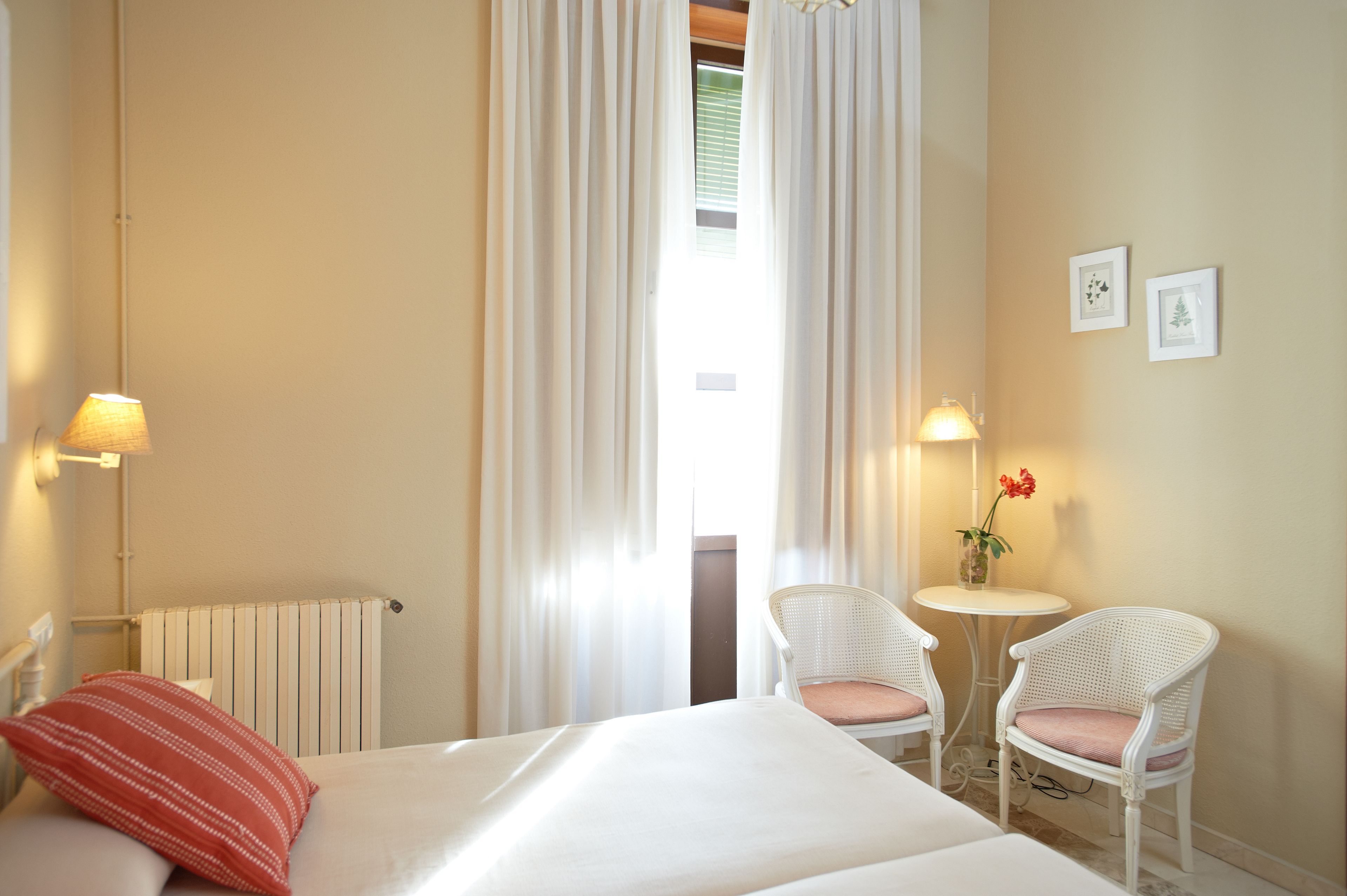 A hotel room with a bed, two white wicker chairs, a small table with a vase of flowers, and two wall-mounted lamps.