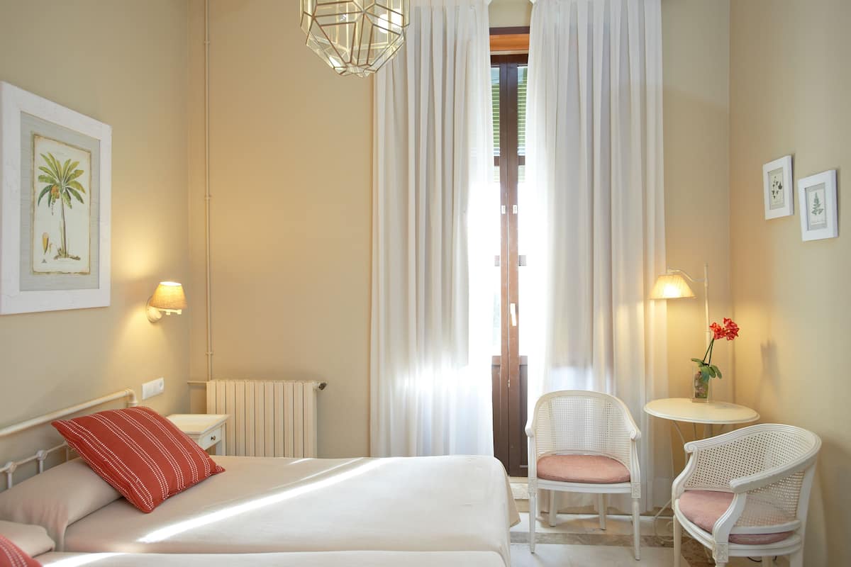 Quarto de hotel com cama, duas cadeiras, uma mesinha, abajur e janela com cortinas.