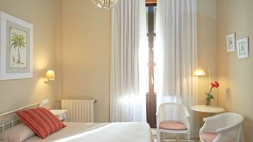 Habitación de hotel con cama, dos sillas, una mesita, lámpara y ventana con cortinas.