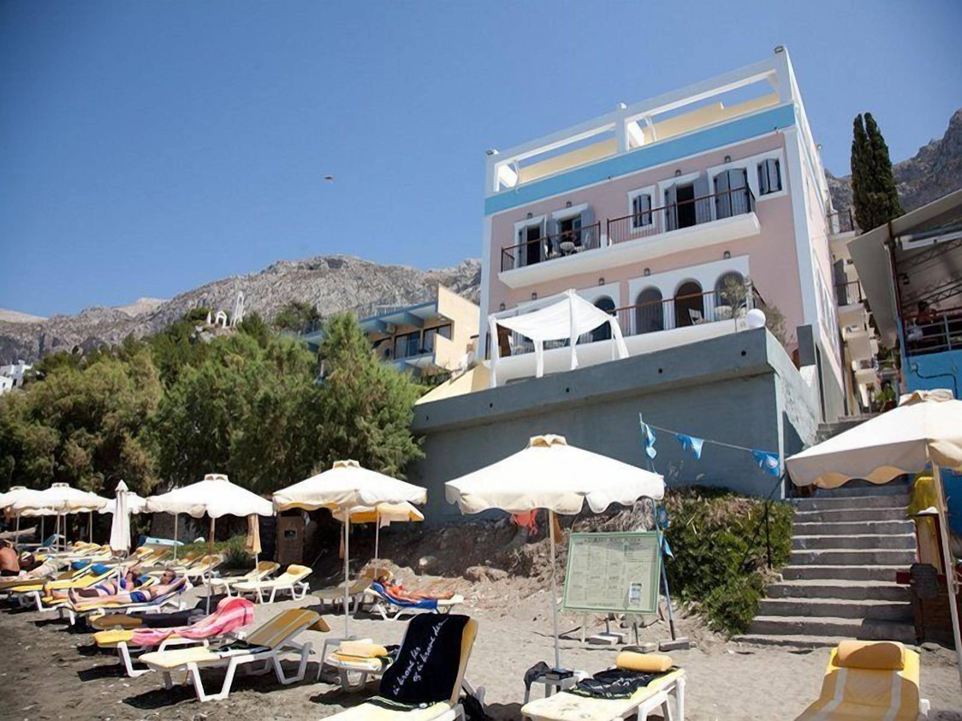 A beachside building with multiple balconies, a sandy beach with sun loungers, and a mountainous backdrop.