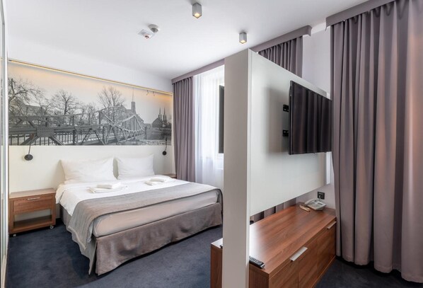 A hotel room with a bed, a bedside table, a desk with a chair, a flat-screen TV, and a large window with curtains.