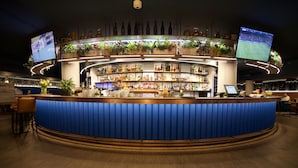 Un espace bar doté d’un comptoir incurvé, de plusieurs étagères remplies de bouteilles et de deux écrans diffusant des événements sportifs.