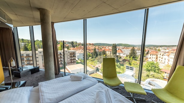 Une chambre d’hôtel avec deux lits, une grande fenêtre donnant sur la ville et un fauteuil vert.