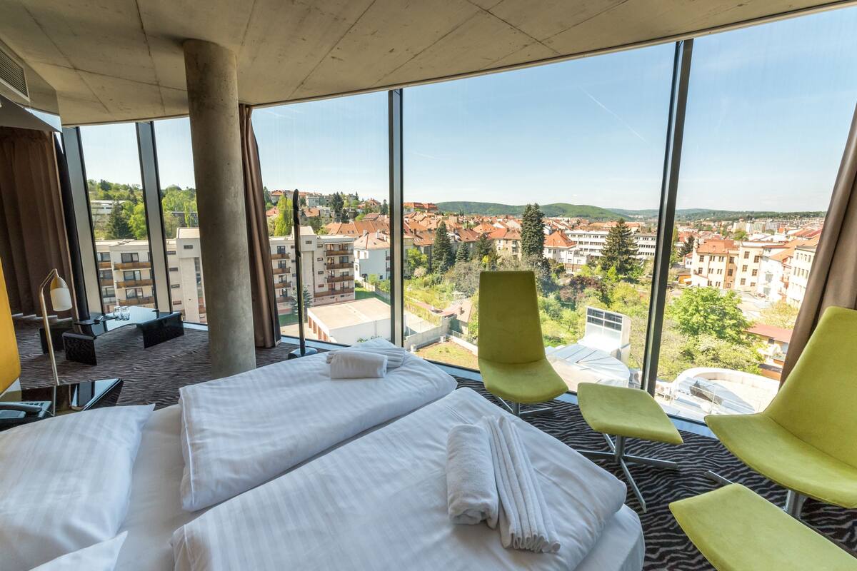 Una habitación de hotel con dos camas, un ventanal amplio que ofrece vistas a la ciudad y una silla verde.