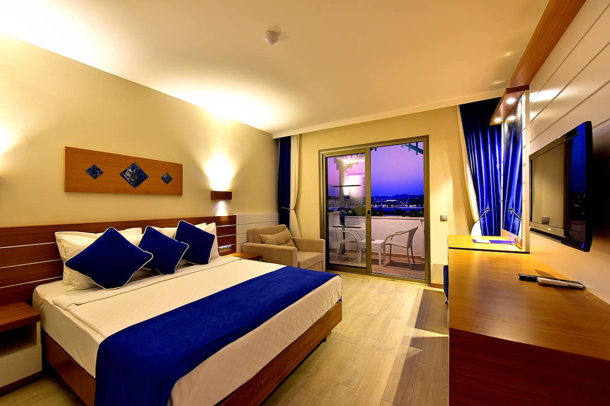 A hotel room with a large bed, a TV, a desk, and a balcony with a city view.