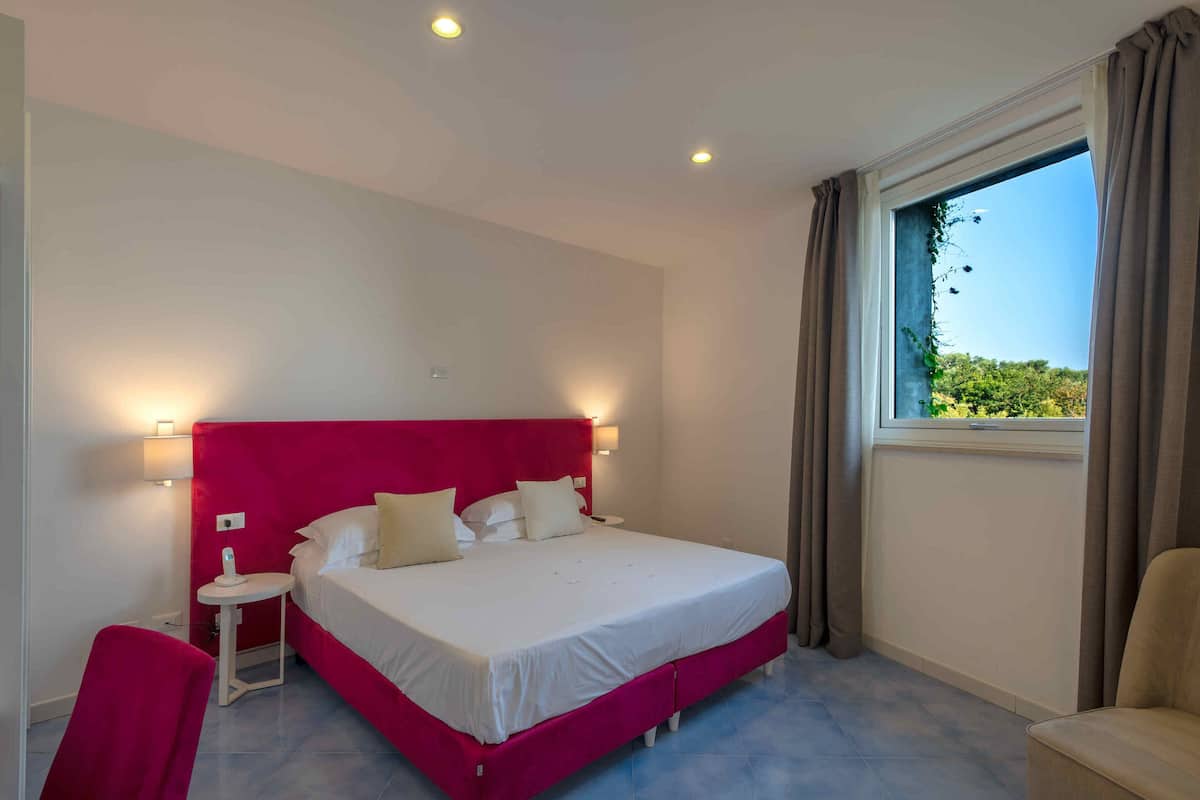 Un dormitorio con una cama grande, una mesita de noche pequeña, una silla y una ventana con cortinas.