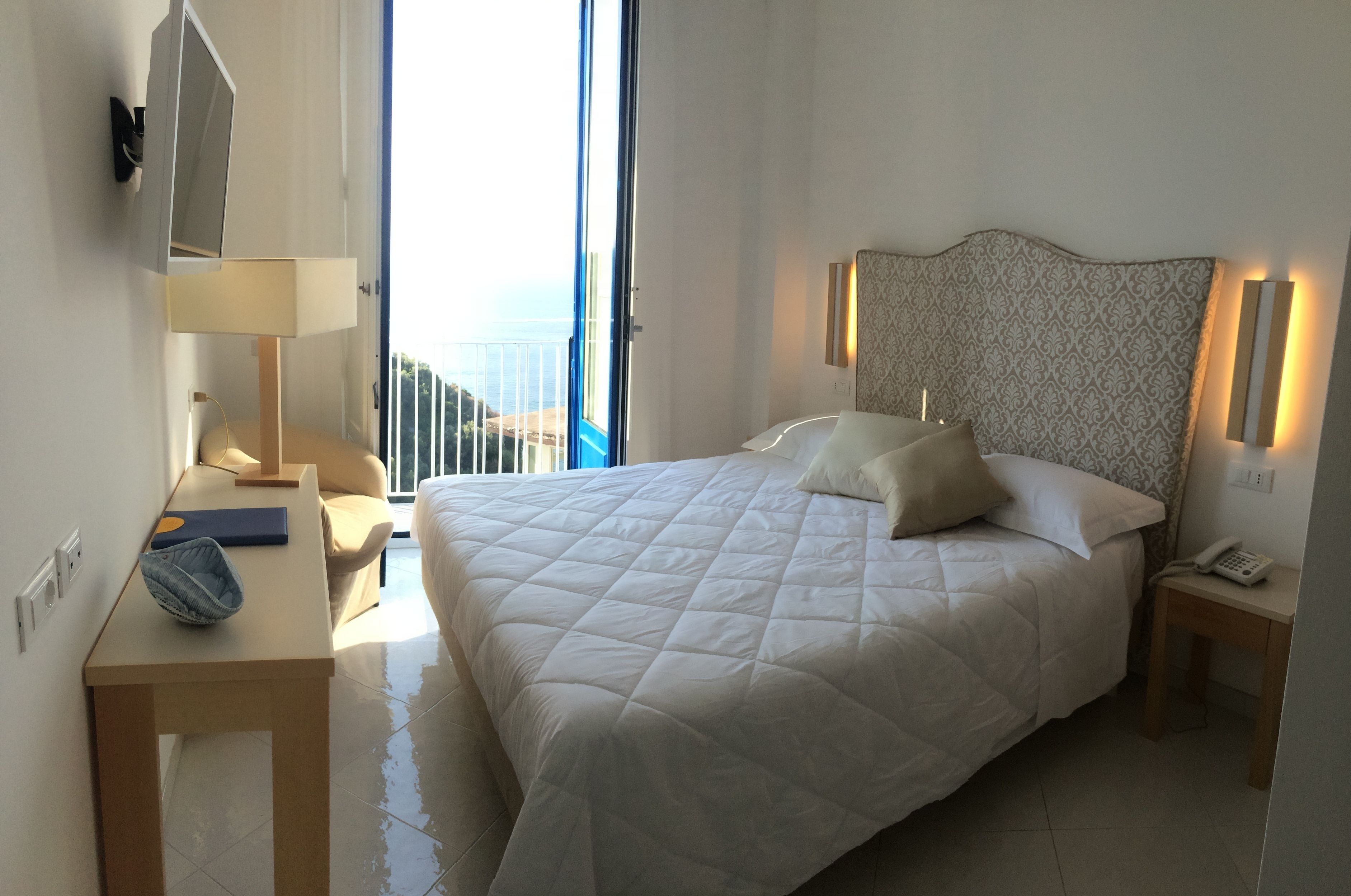 Quarto de hotel com uma cama grande, mesa de cabeceira, abajur, cadeira e vista para o mar.