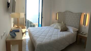 Quarto de hotel com uma cama grande, mesa de cabeceira, abajur, cadeira e vista para o mar.