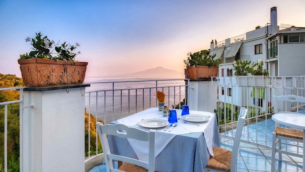 Une terrasse avec une table dressée pour deux personnes, offrant une vue panoramique sur la mer et une montagne lointaine.