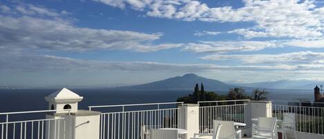 A terrace with white chairs and a table, overlooking a body of water and a mountain in the distance.