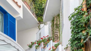 Un escalier étroit aux murs blancs et aux volets bleus, menant à un balcon du deuxième étage, orné d’une balustrade blanche et de plantes en pot.