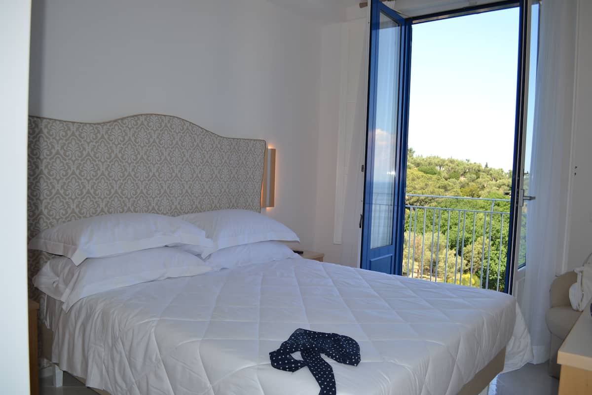 Un dormitorio con cama, una mesita de noche, una ventana que ofrece vista a follaje y una puerta.