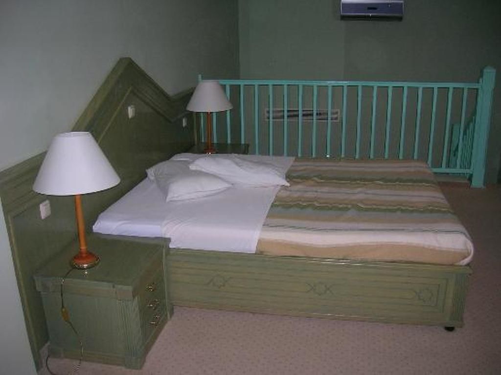 A single bed with a green headboard and footboard, two bedside lamps, and a small nightstand.
