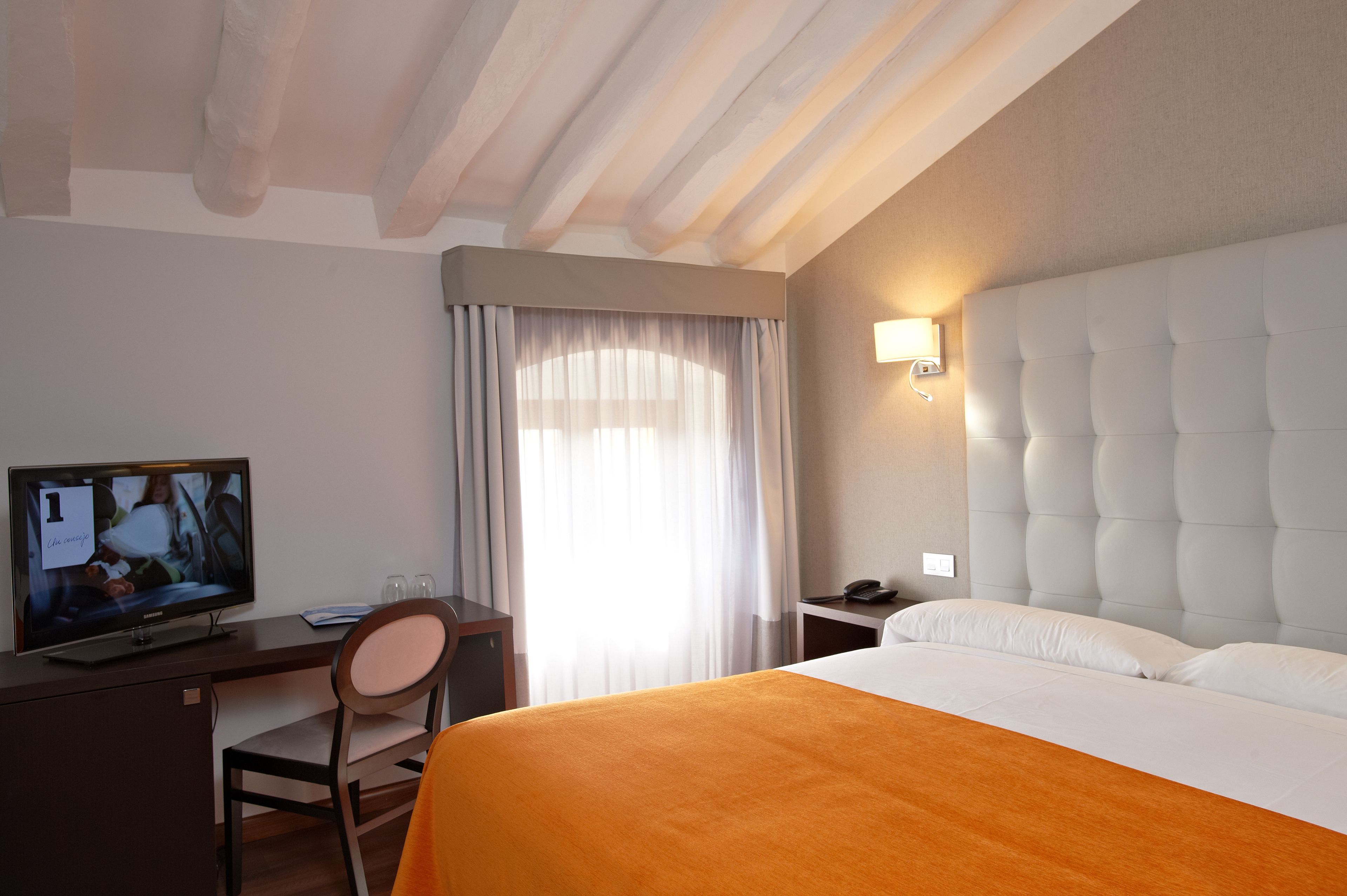A hotel room with a bed, a desk with a television, a chair, a bedside table with a phone, and a window with curtains.