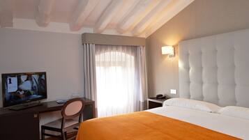 A hotel room with a bed, a desk with a television, a chair, a bedside table with a phone, and a window with curtains.