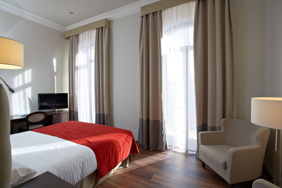 A hotel room with a bed, a desk with a television, a chair, and large windows with curtains.