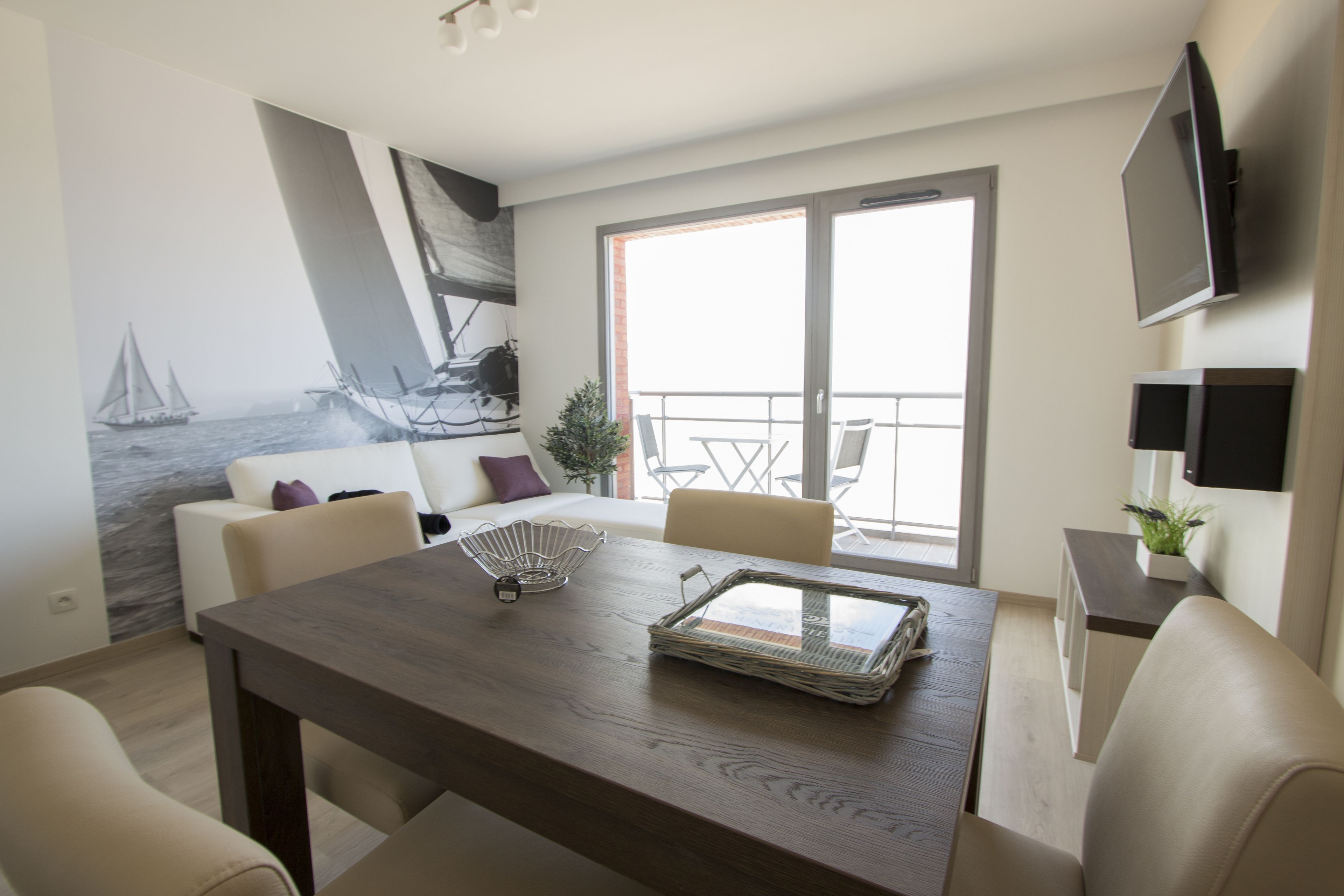 A modern living room with a dining table, sofas, a TV, and a large window with a view of the sea.